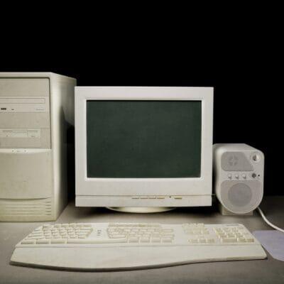 An old-school personal computer with a box-shaped monitor and a white keyboard. There is also a bulky speaker and mouse.