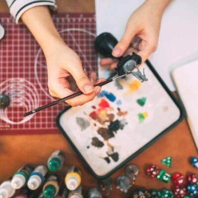 A person hand-painting a small gray tabletop figurine. The table has paints, dice, and other figures on it.