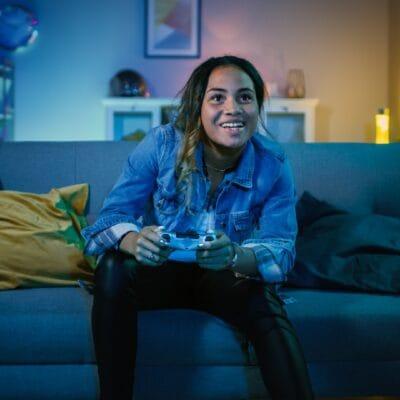 A woman with messy hair leans forward on her couch, playing a video game. the lights around her are soft and blue.