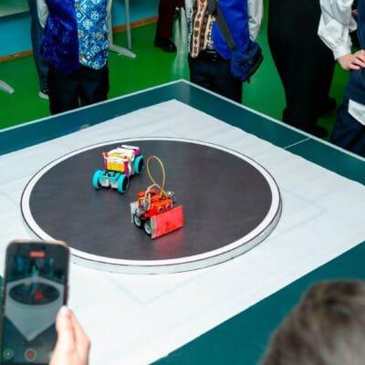 A group of students gathered around a small lifted arena. In the center of the arena, two small robots interact.