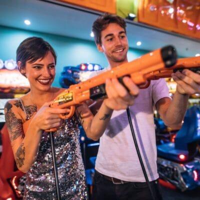 A pair of smiling arcade visitors standing side-by-side as they aim orange light guns toward a game screen.