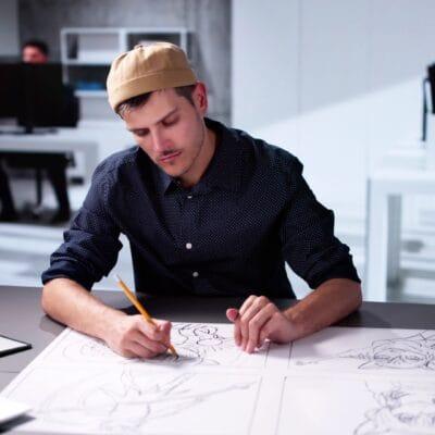 A comic artist sketching out panels on a large sheet of white paper while sitting in a grey-and-white office space.