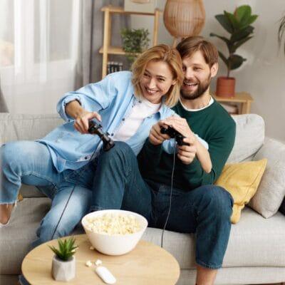 Happy couple sits on a sofa playing video games with controllers, popcorn bowl on table, and green plants in the background.
