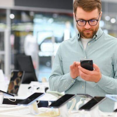 A well-dressed man is in an electronics store looking at cell phones that are for sale. He holds one in his hands.