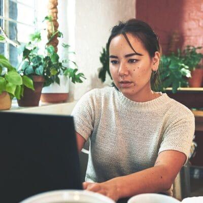 A young person sitting in their studio workspace and looking focused as they type on a large black laptop on a table.