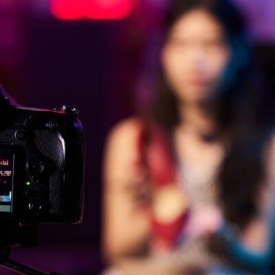 A digital content creator sits in front of a camera placed on a tripod. The lighting is low with a red, purple cast.