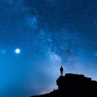 The silhouette of a single person standing on a rocky ledge in front of a deep blue sky filled with stars.