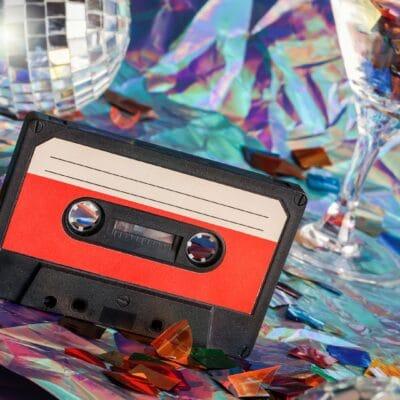 A black-and-red cassette player sitting on a surface covered in iridescent confetti alongside two disco balls and a glass.
