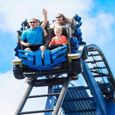 A man in a blue shirt, wearing sunglasses, sits next to a boy wearing an orange polo on a roller coaster.