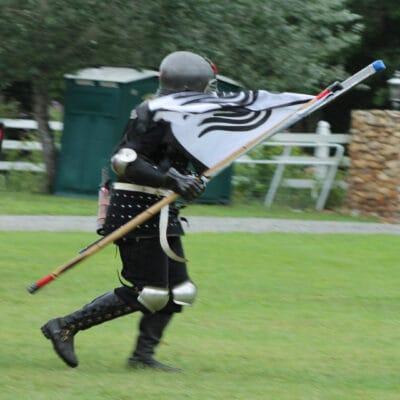 Two people, dressed as medieval knights, run across a grassy area. One holds a flag, and the other has a shield.
