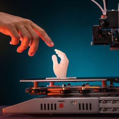 A close-up of a hand about to touch a 3D printed small white hand placed on top of a 3D printing machine.