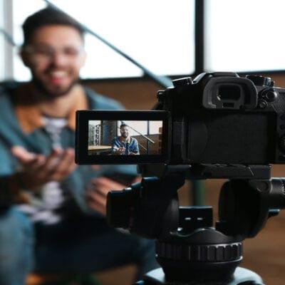 A DSLR camera on a tripod films a smiling man gesturing in an indoor setting, with the focus on the camera screen.