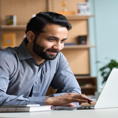 A smiling man sits at a white desk looking at a laptop. A book and a cellphone sit next to the laptop.