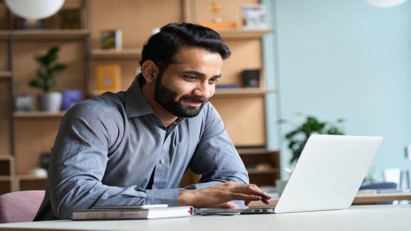 A smiling man sits at a white desk looking at a laptop. A book and a cellphone sit next to the laptop.
