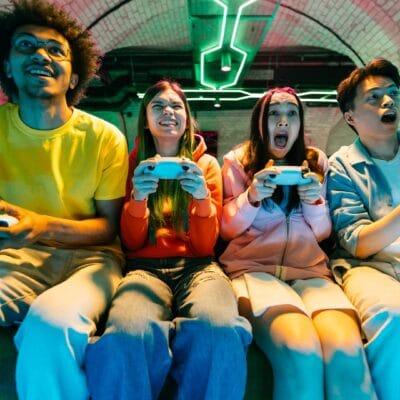 A group of friends sits together on a couch, playing video games. They are in a home game room that features neon lights.