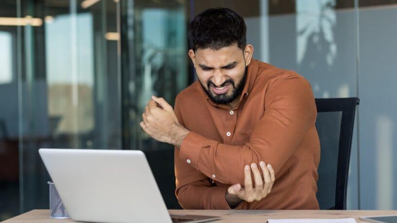 A man wearing a brown dress shirt, sitting behind a desk with a computer on top while holding his elbow in pain.