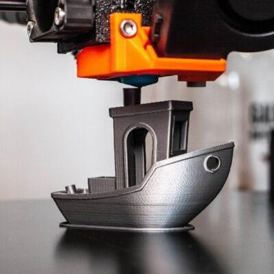 A 3D printing machine with an orange tip, floating on top of a grey 3D printed boat on top of a shiny surface.