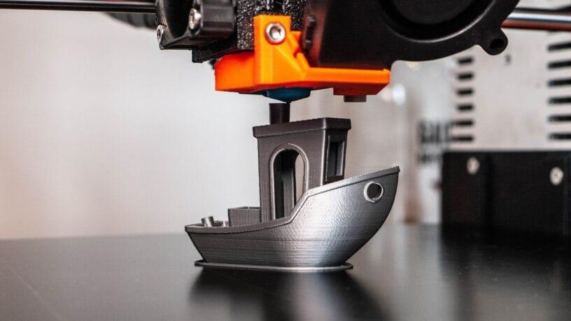 A 3D printing machine with an orange tip, floating on top of a grey 3D printed boat on top of a shiny surface.