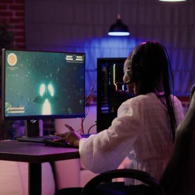 A woman sitting at her gaming setup on her desk. There is a computer tower and a game playing on the screen.