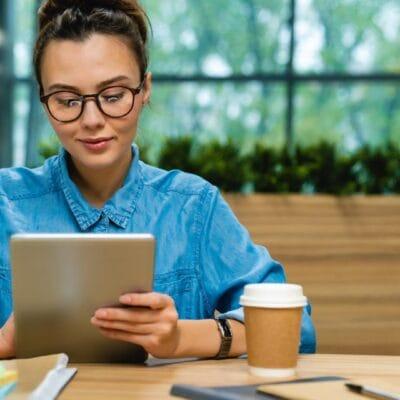 A woman sits at a wooden table looking at an iPad. A to-go coffee cup, a notepad, a pen and a clipboard sit on the table.