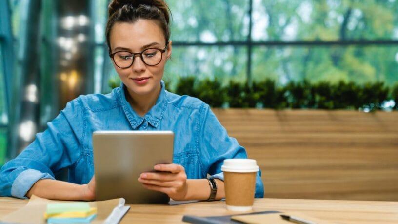 A woman sits at a wooden table looking at an iPad. A to-go coffee cup, a notepad, a pen and a clipboard sit on the table.