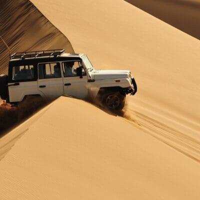 A white off-road SUV drives over steep desert sand dunes, kicking up sand, with a red safety flag mounted on the roof rack.