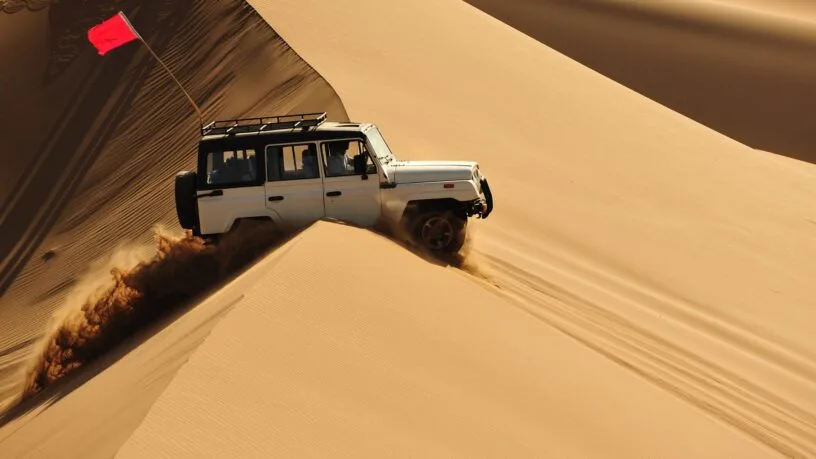 A white off-road SUV drives over steep desert sand dunes, kicking up sand, with a red safety flag mounted on the roof rack.