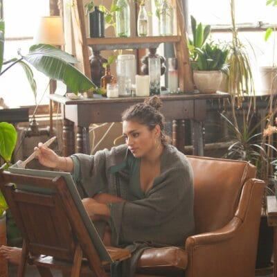 A woman sitting in a chair and painting on a canvas. The room she is in has a lot of green plants scattered around.