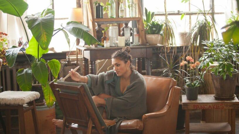 A woman sitting in a chair and painting on a canvas. The room she is in has a lot of green plants scattered around.