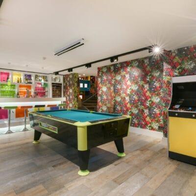 A colorful game room features a pool table, arcade cabinet, and bright bar beneath modern lighting in a basement.