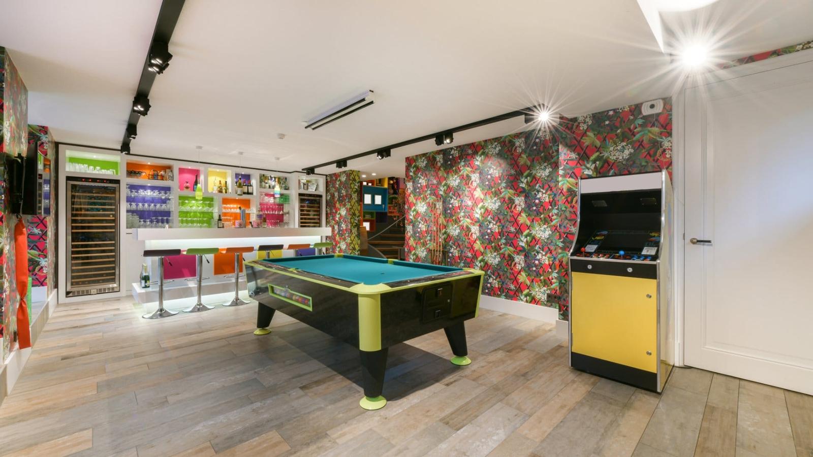A colorful game room features a pool table, arcade cabinet, and bright bar beneath modern lighting in a basement.