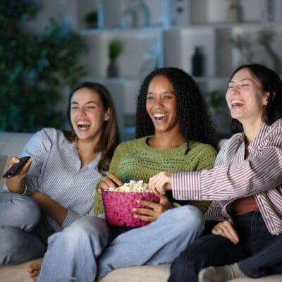 Three women laugh and reach into a bowl of popcorn on a couch at night, their faces lit by the blue glow of a TV screen.