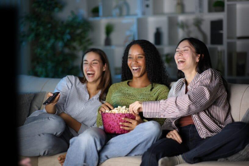 Three women laugh and reach into a bowl of popcorn on a couch at night, their faces lit by the blue glow of a TV screen.