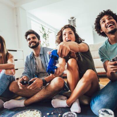 Four friends laugh and play video games together in a bright living room with snacks on the floor nearby.