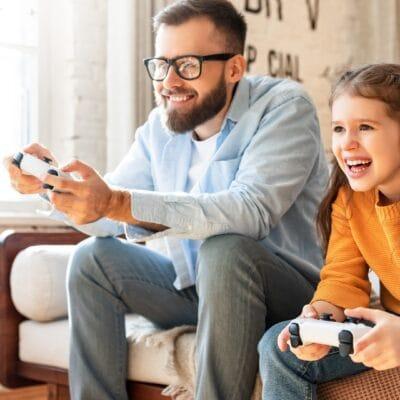 A father and daughter sit on a couch holding white video game controls. They are both smiling and wearing jeans.