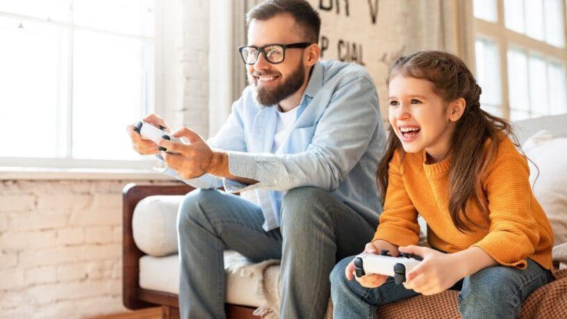 A father and daughter sit on a couch holding white video game controls. They are both smiling and wearing jeans.